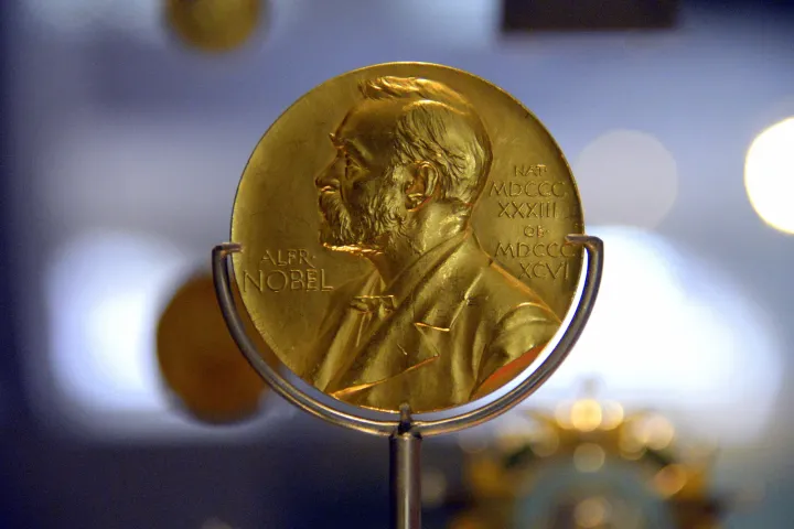 A gold Nobel Prize medal on a stand, showing Alfred Nobel's face in profile, with soft lights glowing behind it
