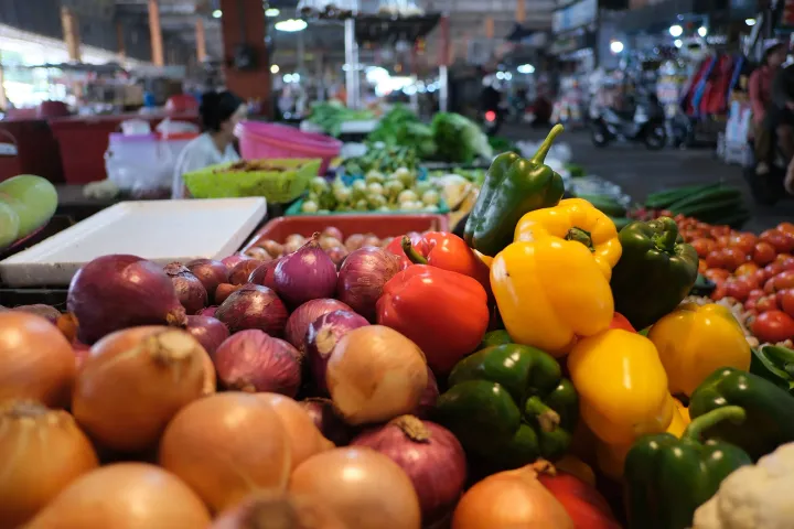 A market stall piled with red, yellow and green bell peppers beside heaps of red and brown onions