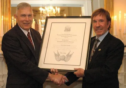 Two men in dark suits shake hands and hold a framed certificate in an ornate room