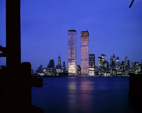 The twin towers of the World Trade Center lit up at dusk across the water, with the New York skyline