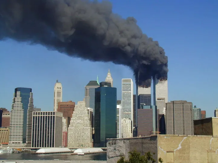 The twin towers of the World Trade Center burning, with thick black smoke pouring across a blue sky above lower Manhattan