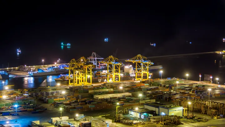 A container port at night: yellow cranes load a ship beside stacks of containers under floodlights