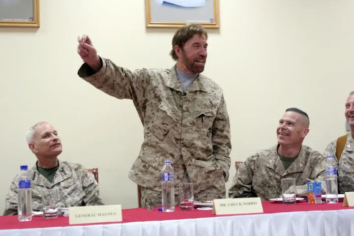 Chuck Norris, in a Marine desert uniform, stands and gestures at a table where Marine officers watch and laugh